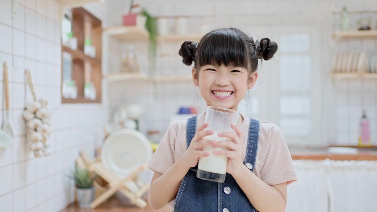 詳情圖-亞洲可愛的小孩拿著一杯牛奶在家里的廚房里喝。學(xué)齡前兒童或女兒被牛奶弄臟后，用手臂擦嘴，然后微笑著看鏡頭。