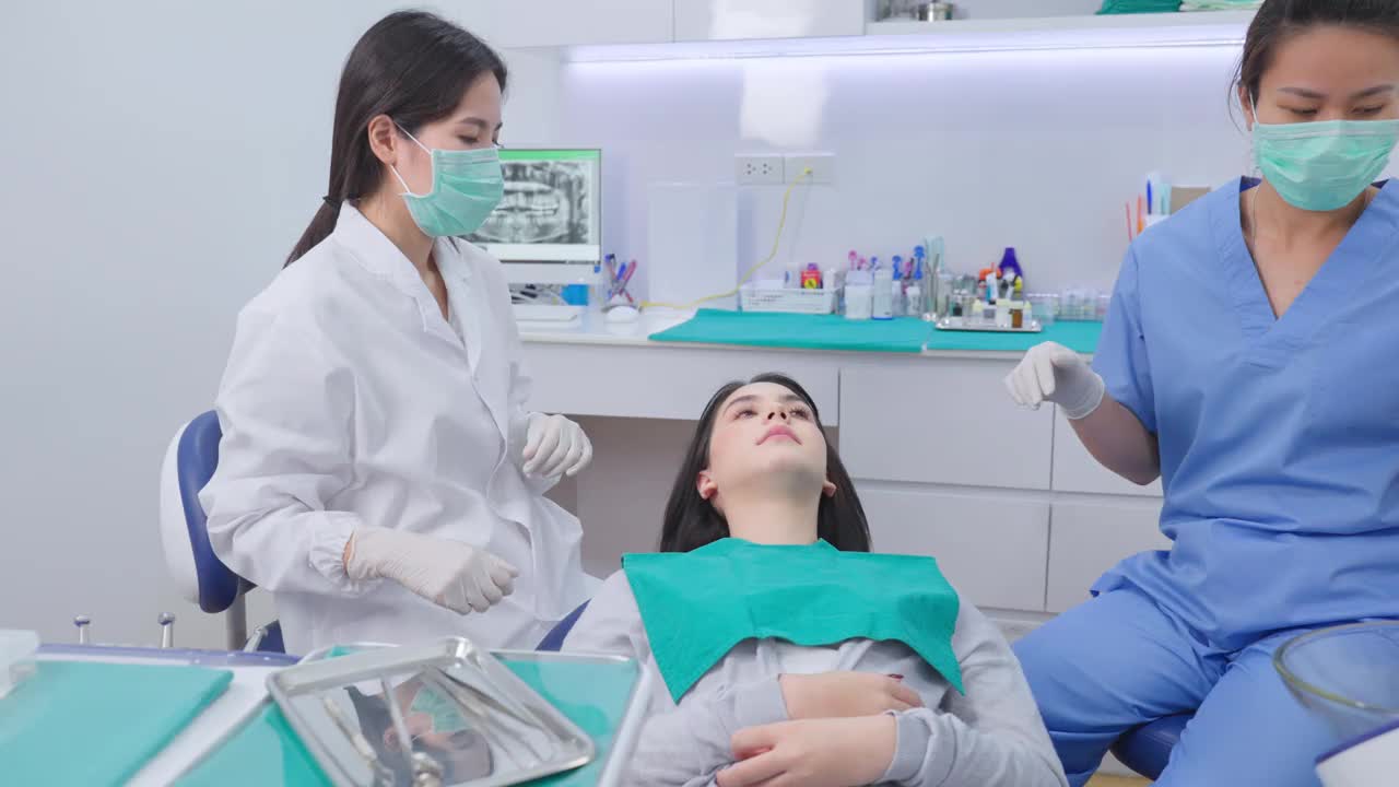 亚洲年青牙医戴上口罩，使用医疗器械进行口腔护理治疗。在诊所治疗过程中，协助坐在吸痰机旁保持患者口腔干燥。