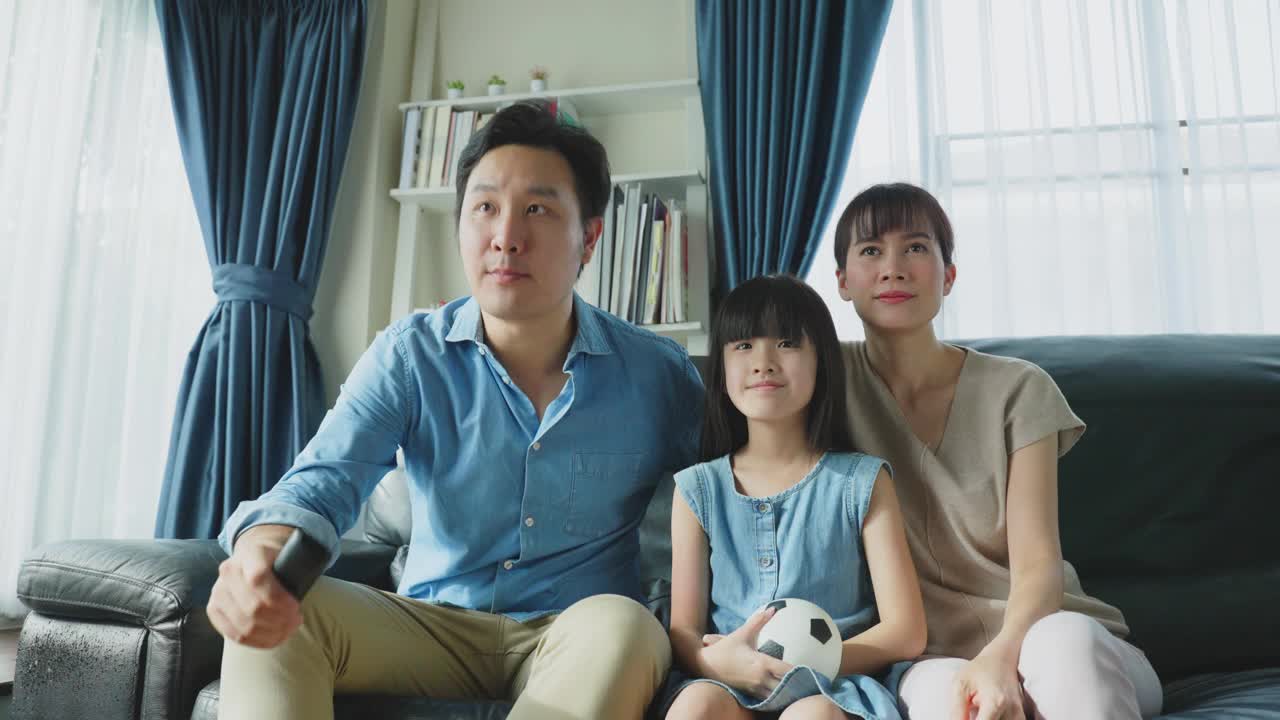 亚洲幸福的父母和女儿坐在家里客厅的沙发上。兴奋的家庭，年轻的小女孩球迷与父母拿着足球看电视体育比赛，庆祝球队的胜利。