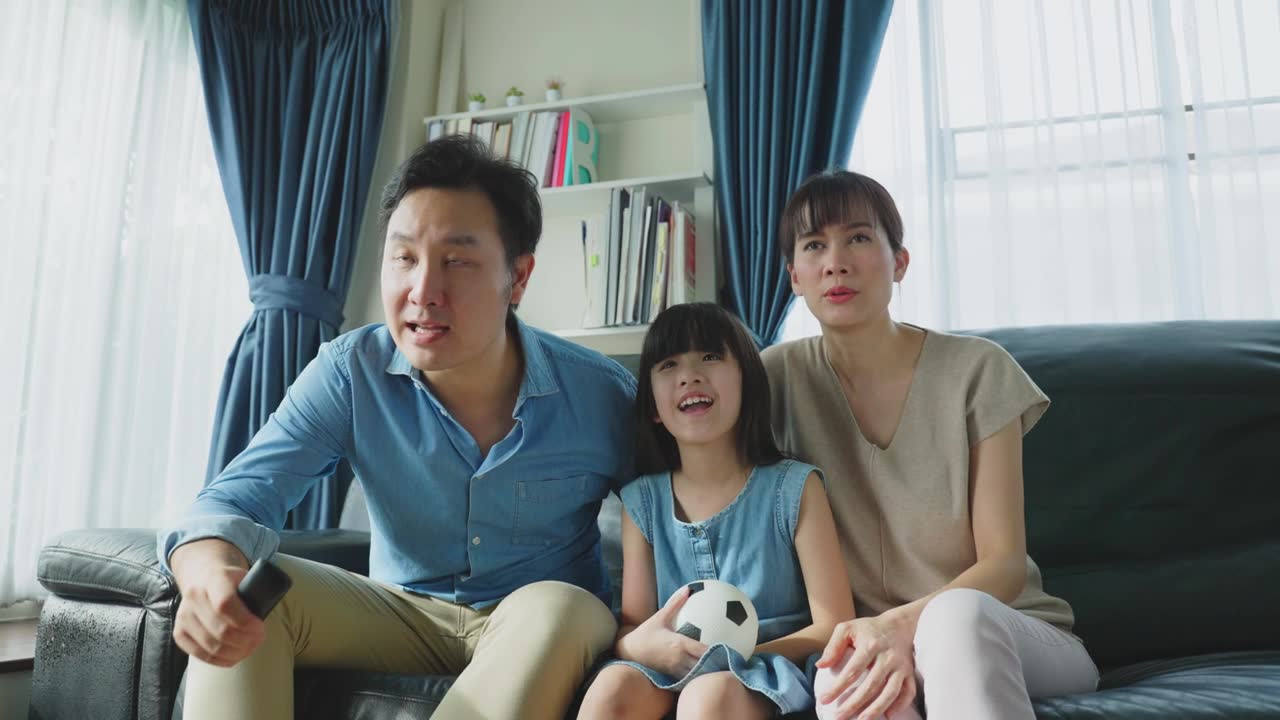 詳情圖-亞洲幸福的父母和女兒坐在家里客廳的沙發(fā)上。興奮的家庭，年輕的小女孩球迷與父母拿著足球看電視體育比賽，慶祝球隊(duì)的勝利。