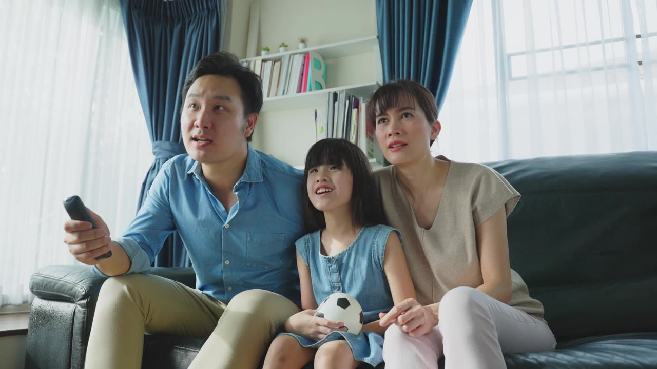 詳情圖-亞洲幸福的父母和女兒坐在家里客廳的沙發(fā)上。興奮的家庭，年輕的小女孩球迷與父母拿著足球看電視體育比賽，慶祝球隊(duì)的勝利。