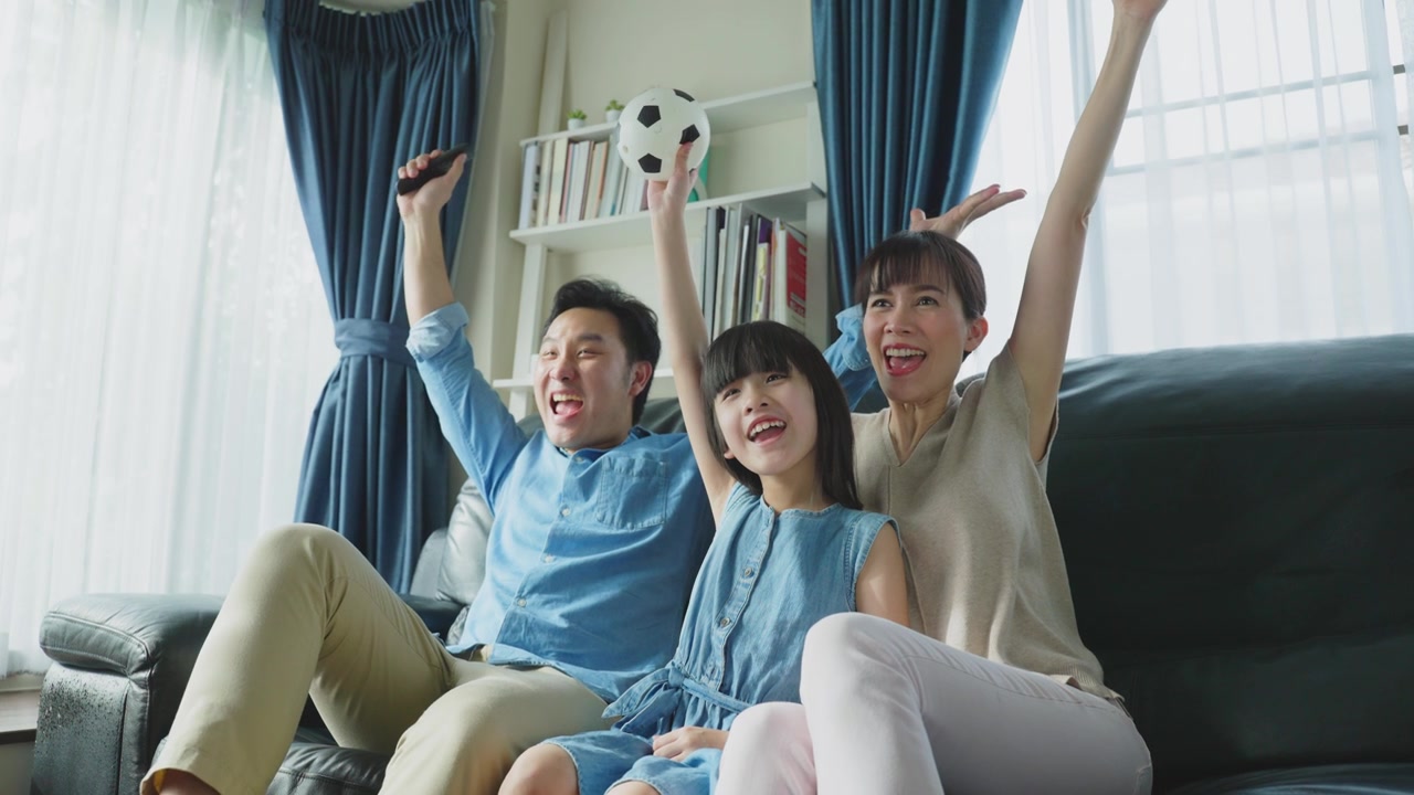 詳情圖-亞洲幸福的父母和女兒坐在家里客廳的沙發(fā)上。興奮的家庭，年輕的小女孩球迷與父母拿著足球看電視體育比賽，慶祝球隊(duì)的勝利。