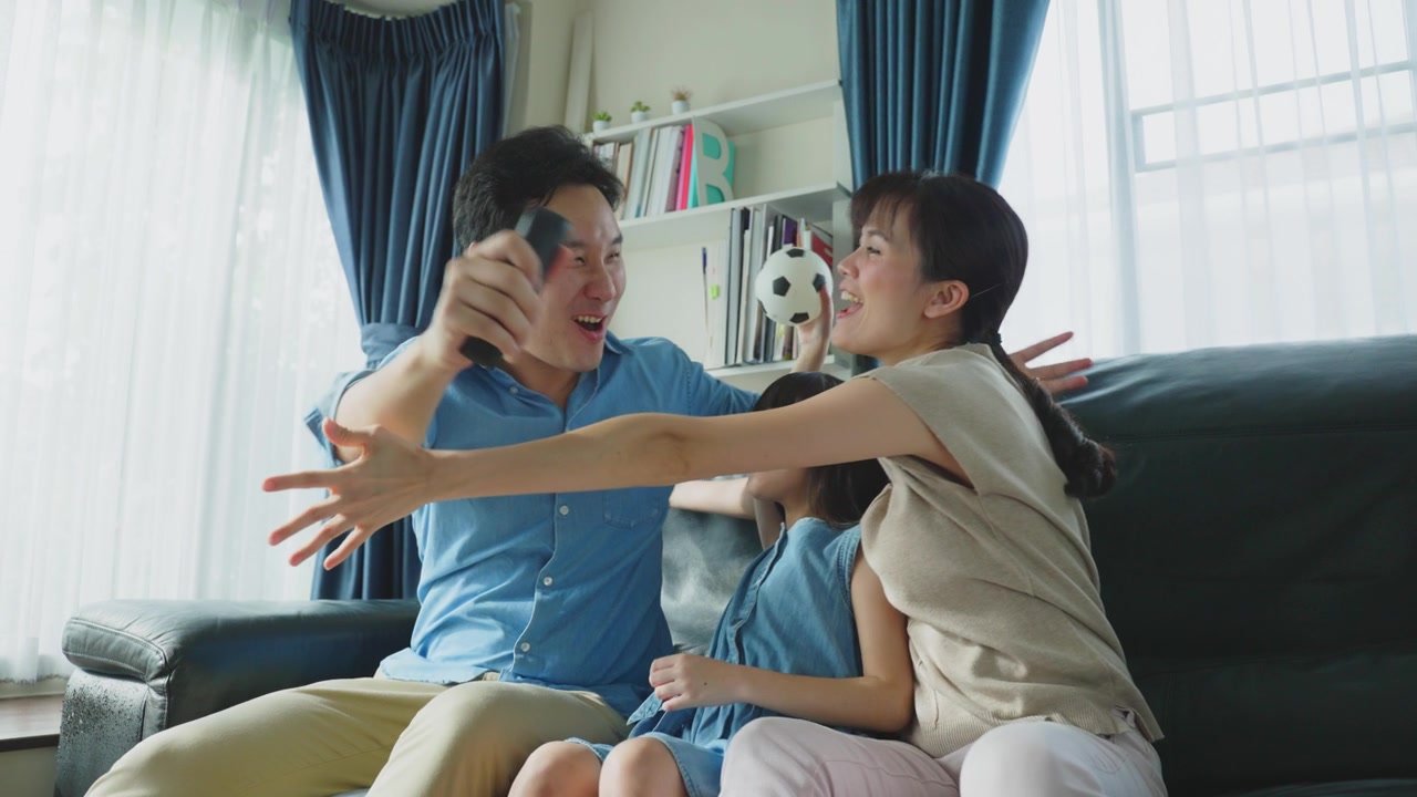 詳情圖-亞洲幸福的父母和女兒坐在家里客廳的沙發(fā)上。興奮的家庭，年輕的小女孩球迷與父母拿著足球看電視體育比賽，慶祝球隊(duì)的勝利。