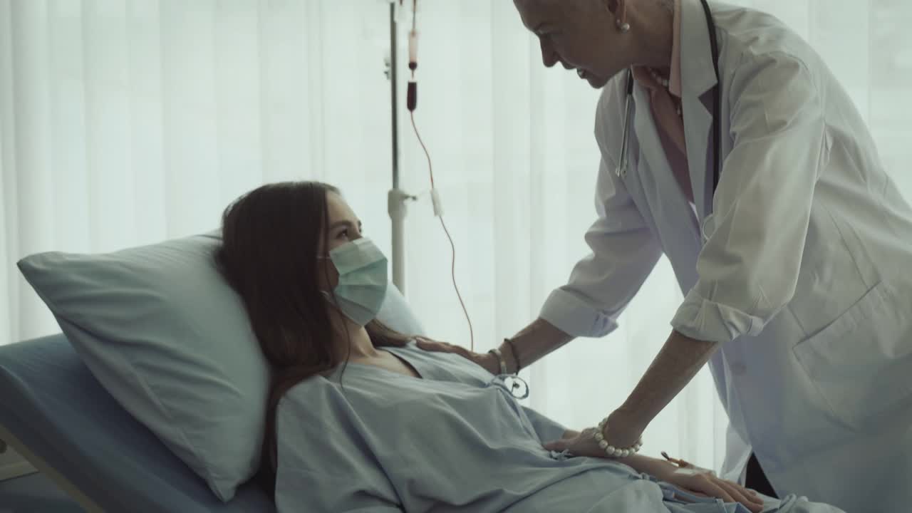 一位女病人躺在病床上，与她的资深医生讨论处方药物的选择。医生回答病人的问题。他们戴着防护口罩。
