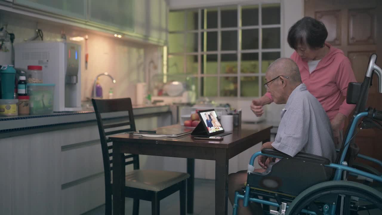 坐在轮椅上的亚洲老人和妻子在晚餐后的闲暇时间在厨房用平板电脑给家人视频通话