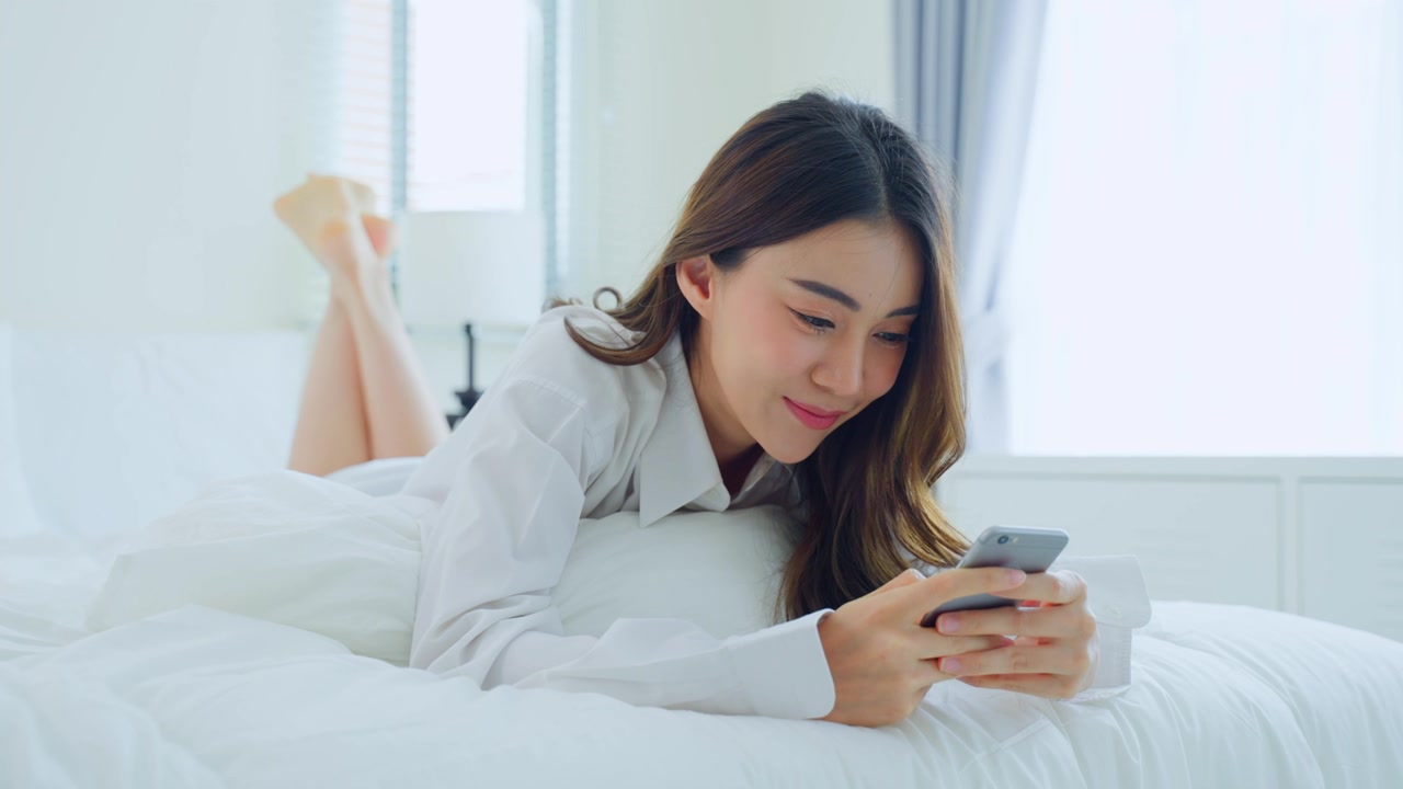 詳情圖-亞洲美女早上在家里的床上用手機(jī)聊天?？鞓?lè)隨意的漂亮女性躺在臥室享受假日周末，在家里用智能手機(jī)和科技交流。