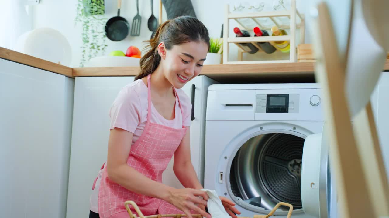 亚洲美女把脏衣服放在家里的洗衣机里。漂亮的女孩穿着围裙坐在地板上，开心地在家里的洗衣机里装衣服。居住房屋保持概念。
