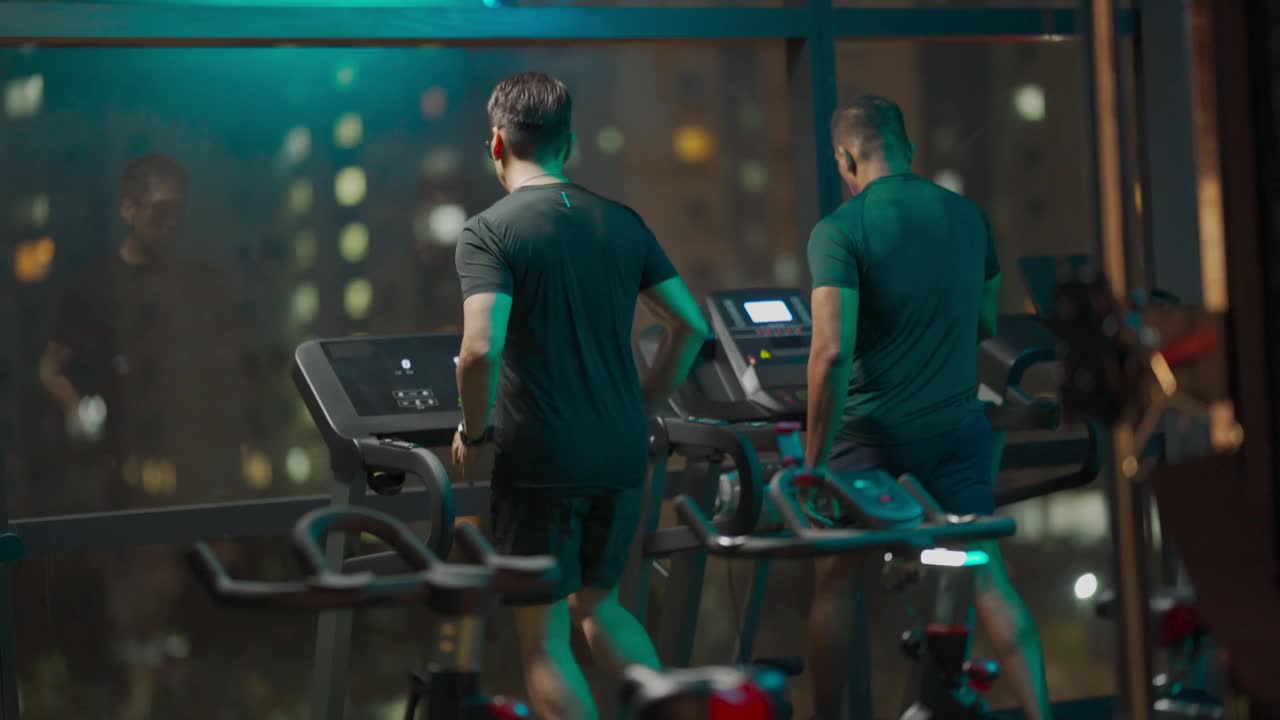 后视图大男子主义的男人力量走跑步机一起在健身房聊天在晚上面对城市的街灯