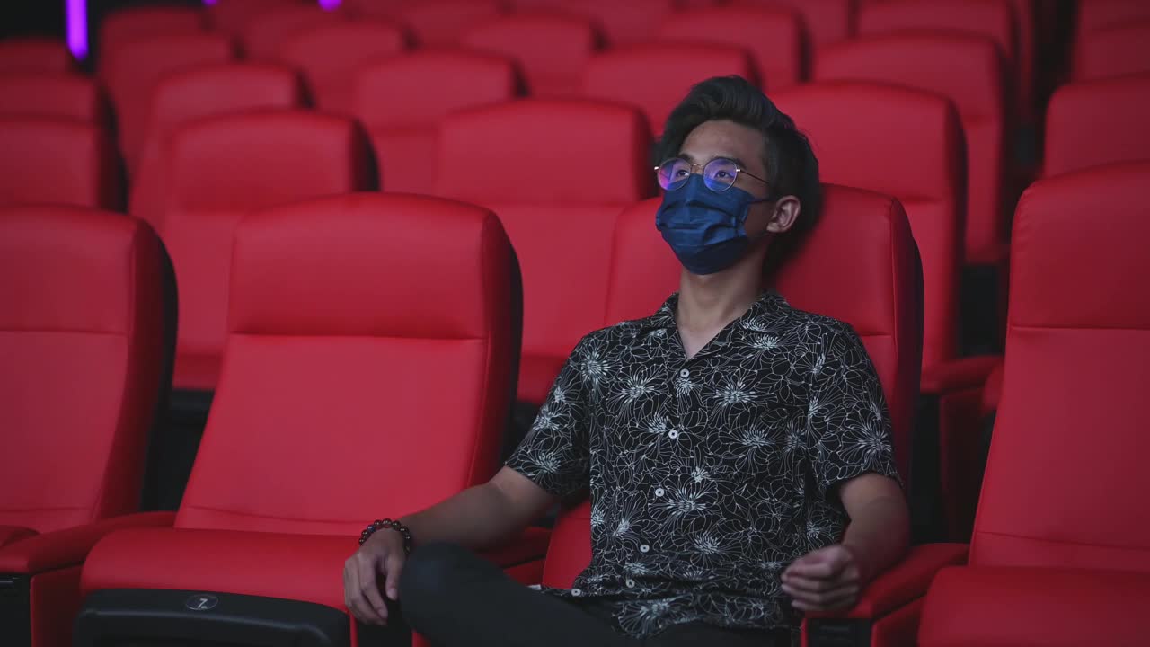亚洲华人少年戴着口罩在电影院看电影，呆若木鸡，跷着二郎腿