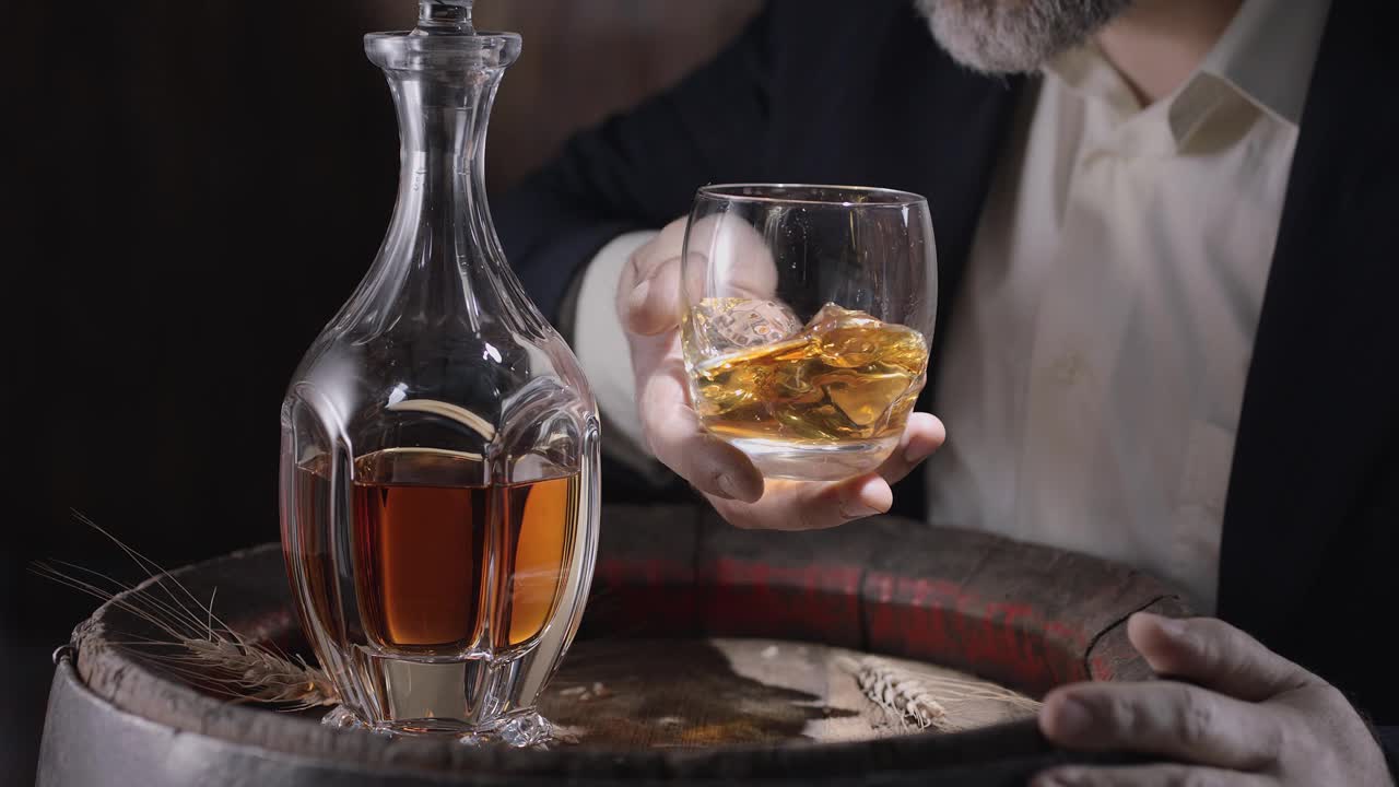 一个穿西装留着胡子的男人(侍酒师)从一个陈年威士忌酒桶里拿出一杯加冰的威士忌，摇了摇酒杯，让威士忌的味道散发出来。慢动作4K, 150 fps，黑魔法大熊Pro G2。