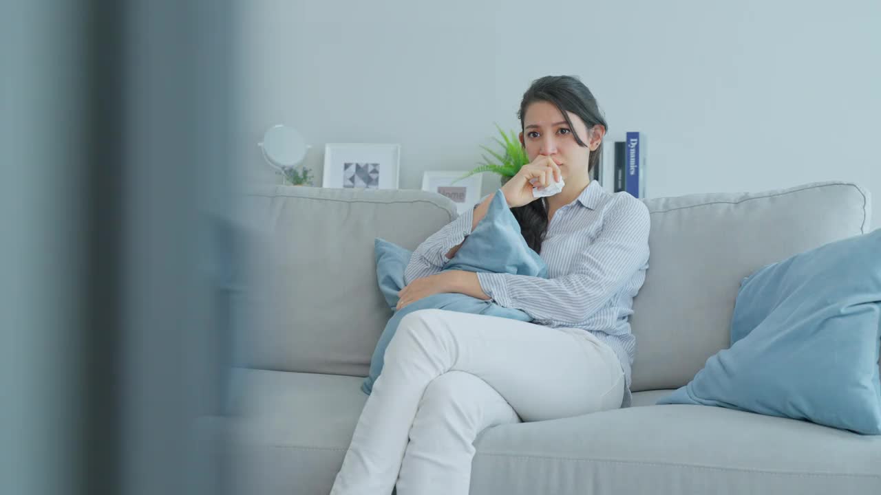 白人爱女人在客厅里看电视上的悲伤电影。漂亮的女性哭着哭着，坐在沙发上用遥控器看电视，在家里看电视上的戏剧和悲伤节目。