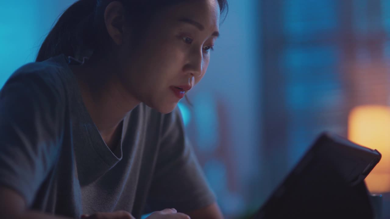 年轻的亚洲女性晚上用数码平板电脑看网络电视