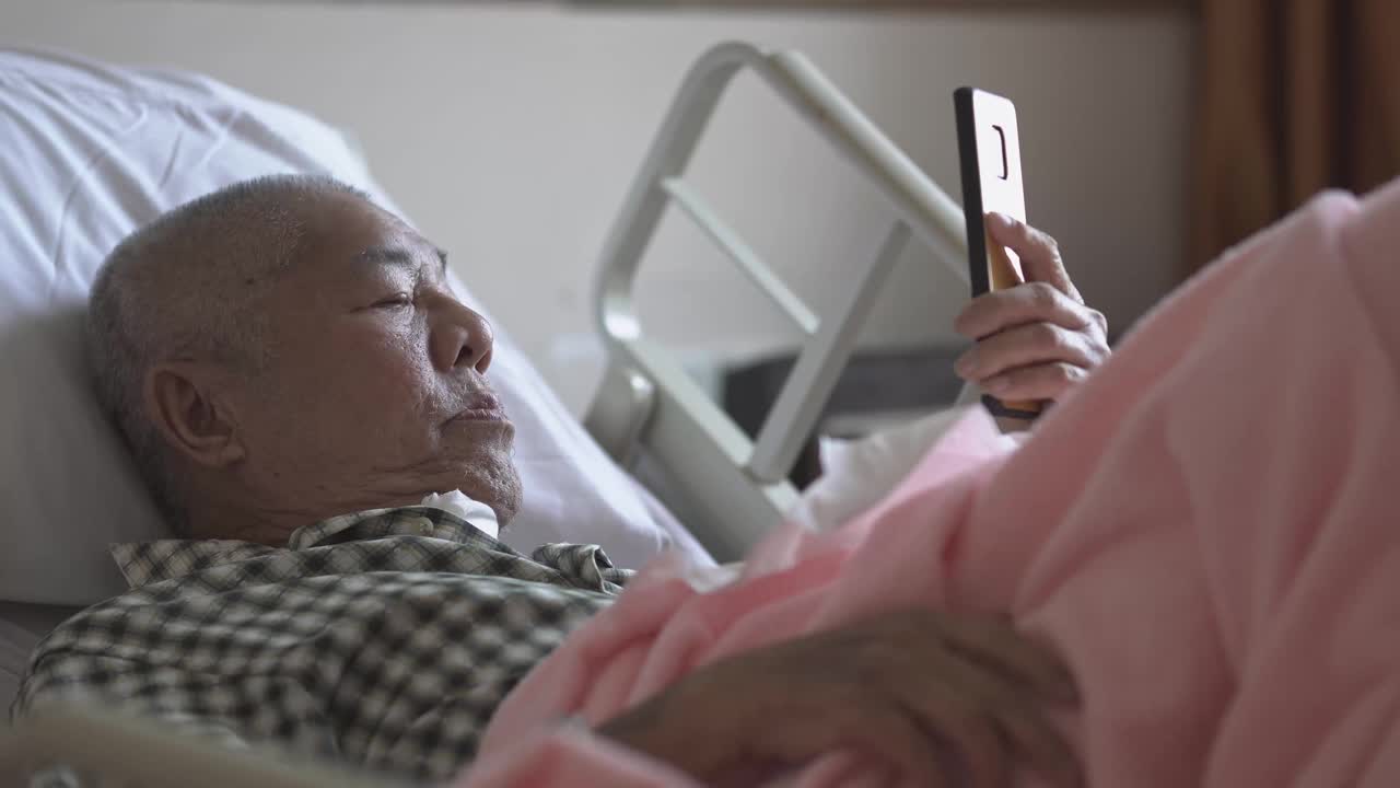 一位亚裔华裔老人躺在医院病房的病床上，用智能手机与家人视频通话