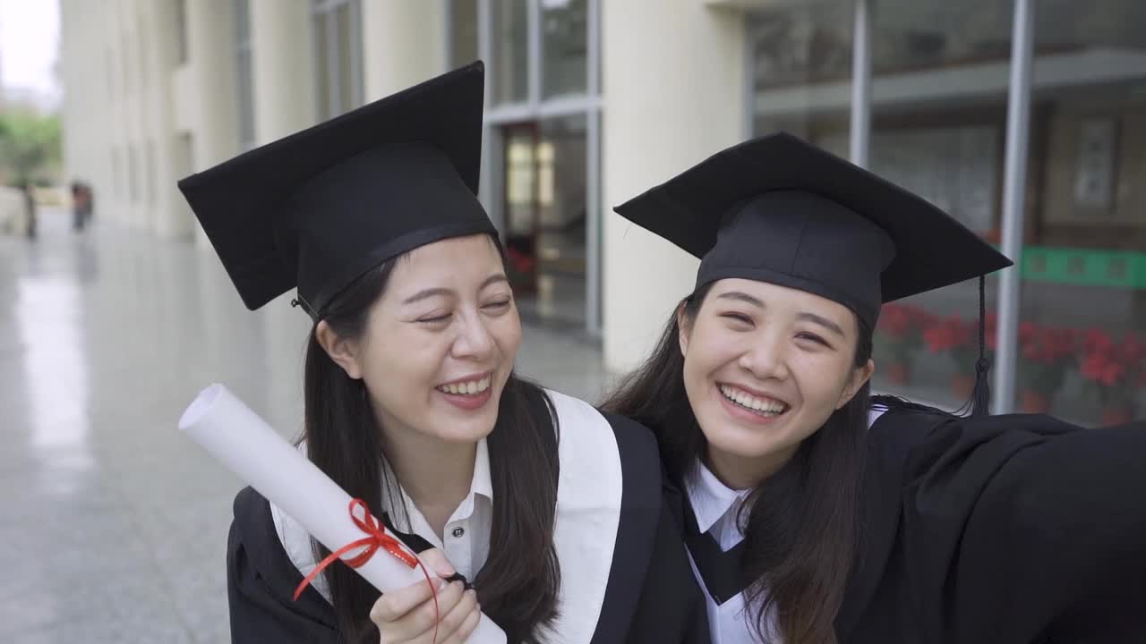 两个亚洲女大学生最好的朋友拥抱在一起，站在户外的草地上，面对镜头竖起大拇指，展示毕业典礼的毕业证书