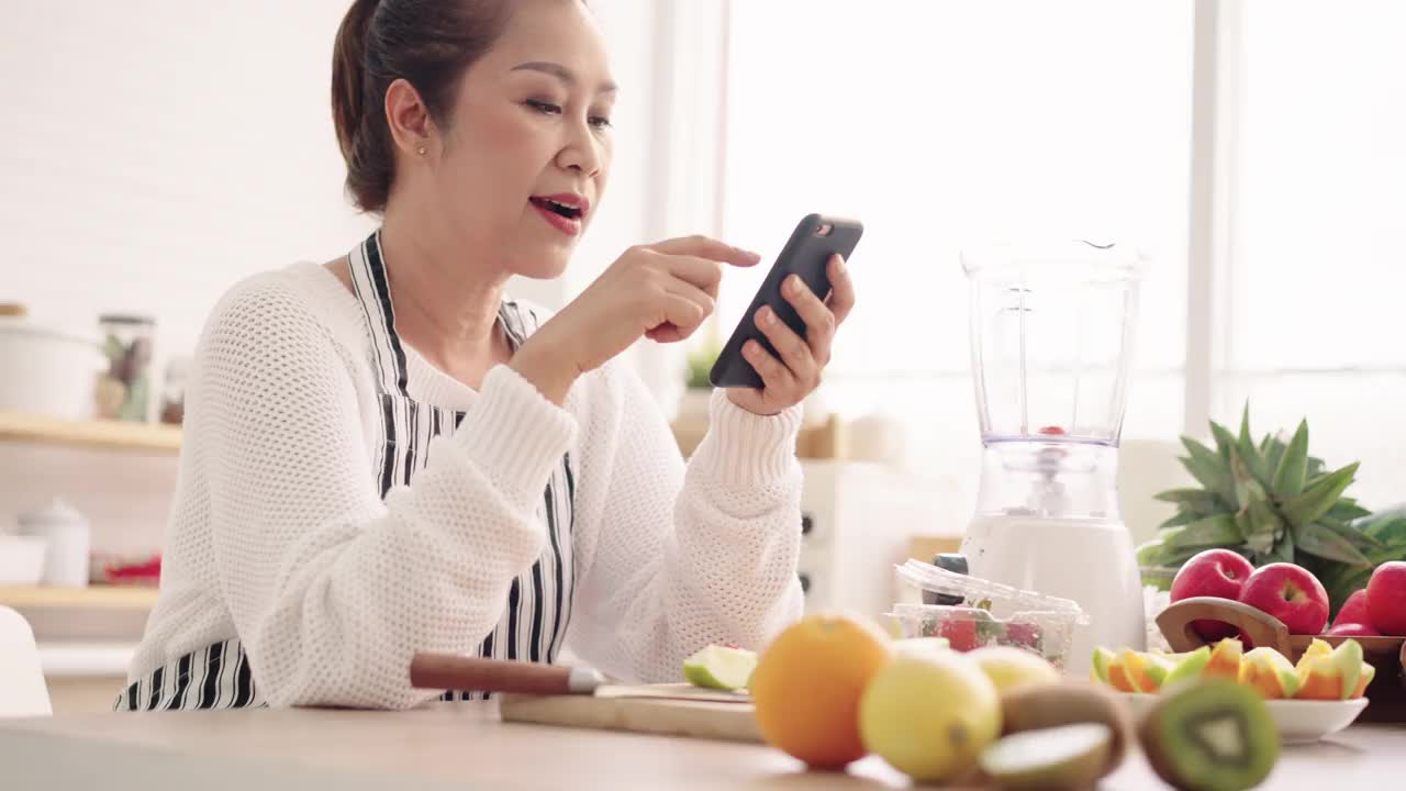 亚洲资深女性一边吃水果一边用手机感到幸福，准备用不同的口味和酸奶自制水果冰沙。健康天然益生菌调味饮料，在厨房学习使用科技