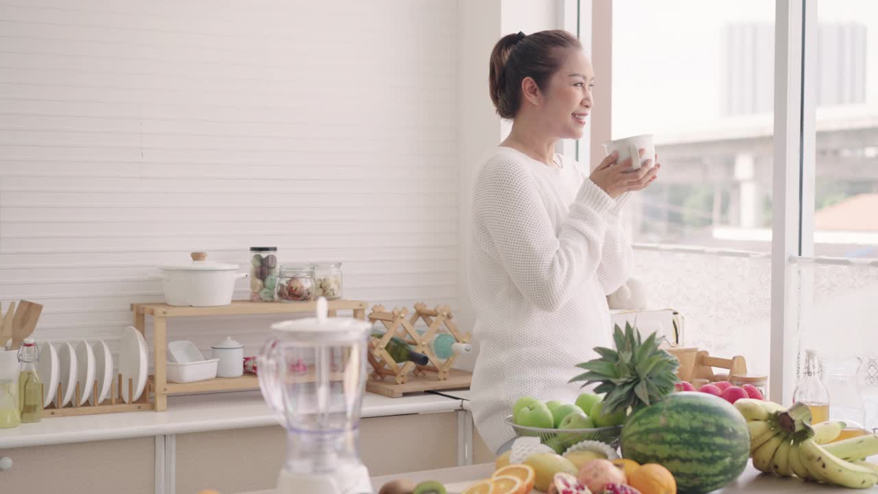 亚洲老年妇女在家里的厨房里喝热牛奶，中间夹着热带水果，西瓜，香蕉，木瓜，青苹果，橙子，用来制作水果奶昔，老年妇女概念退休，活跃的人