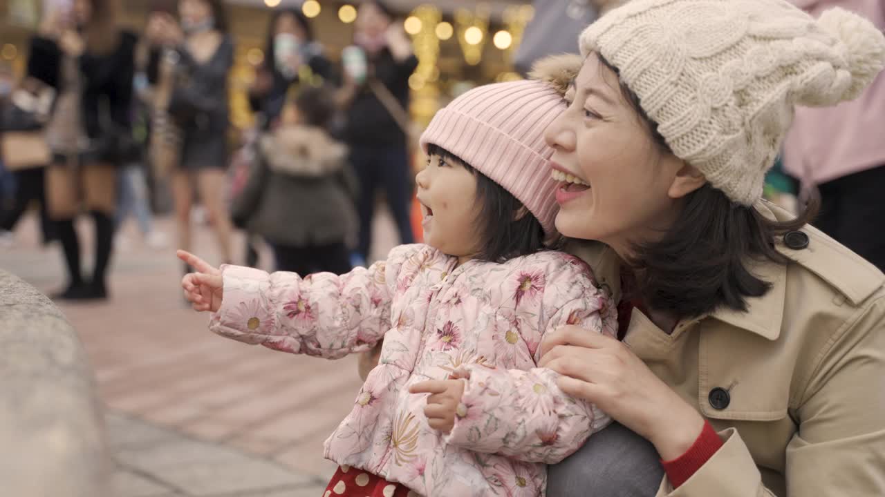 中国台湾的母亲和婴儿笑着看着城市广场上跳舞的喷泉，以一群游客为背景拍照