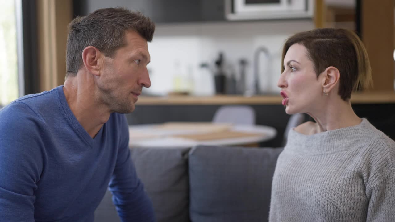 侧视图愤怒的好斗的夫妇尖叫，手势，争吵在家里室内。成年白人男人和女人大喊大叫，互相责骂，争吵。刺激和冲突。