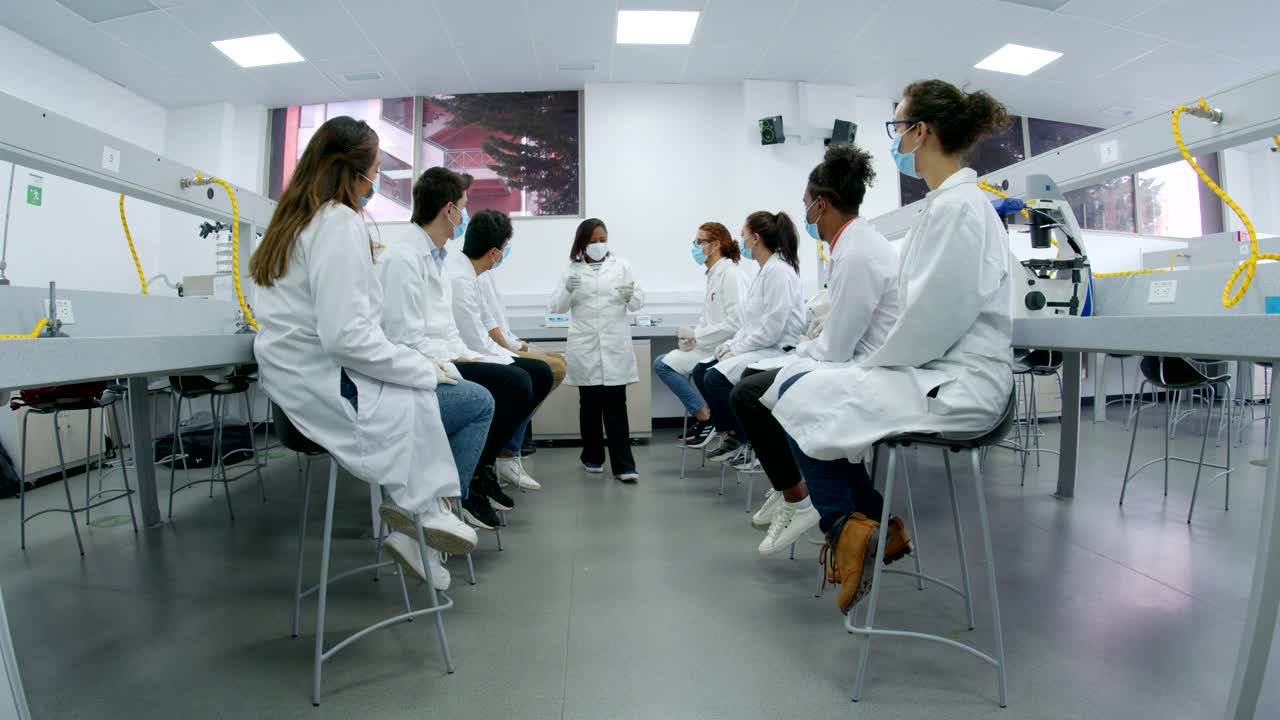 一位非裔美国科学教授在实验室对一群医学院学生讲话，并向他们展示装在两个烧杯里的样本