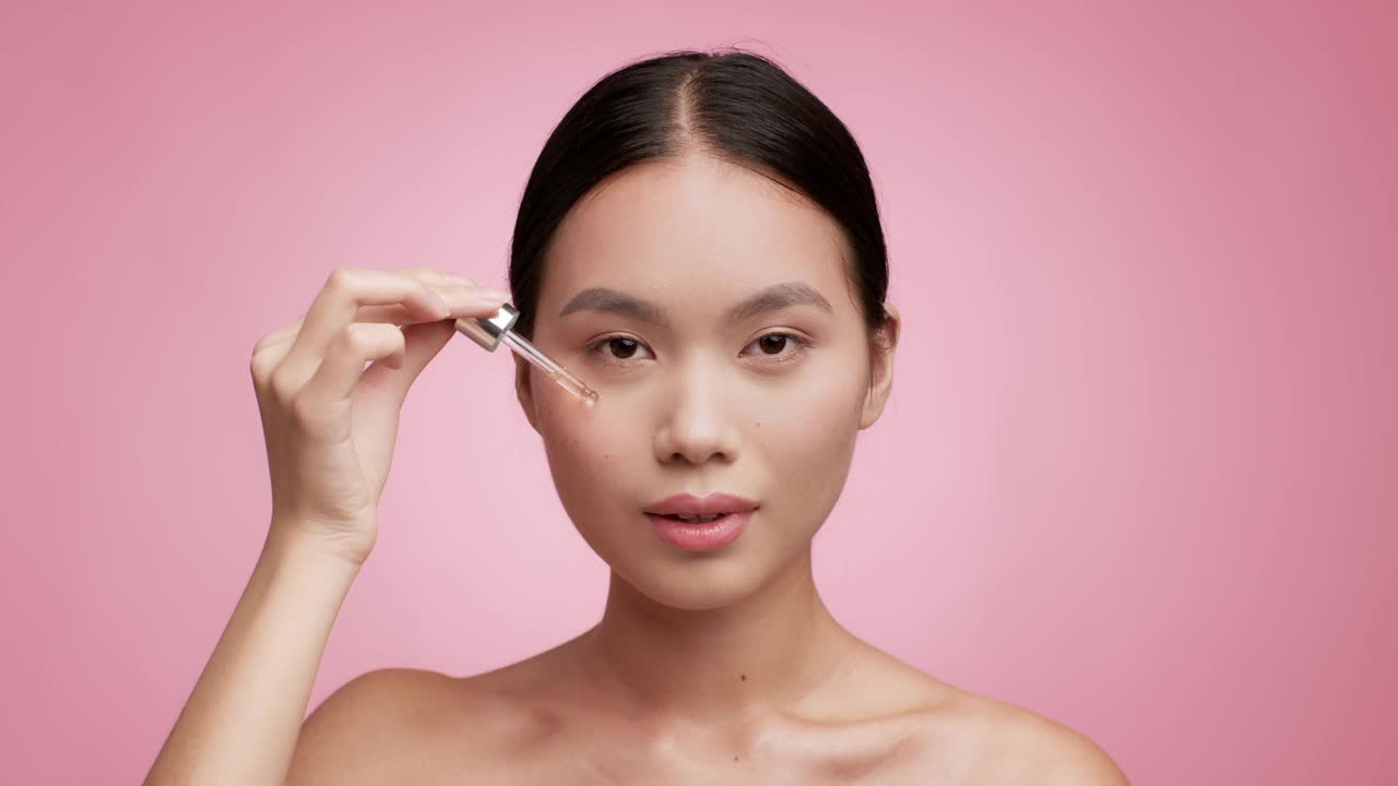 亚洲女性使用滴管在粉红色背景上使用面部精华液