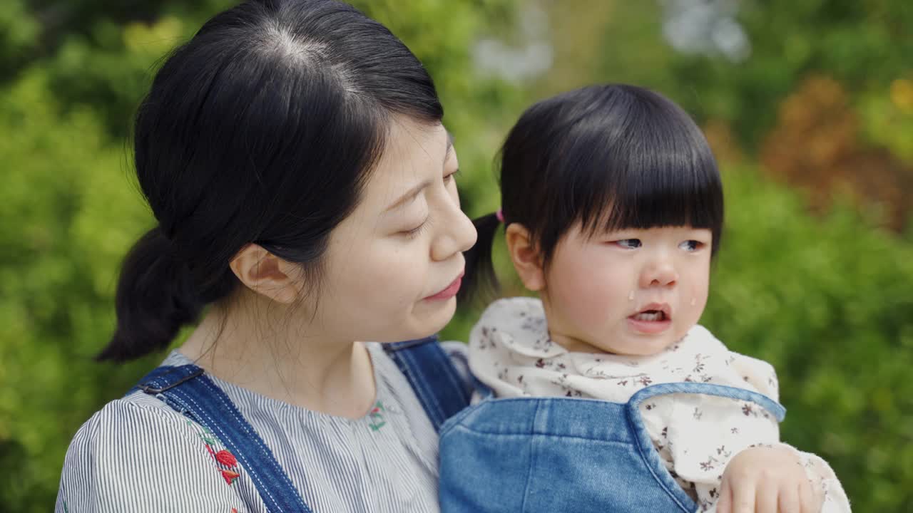 在一个阳光明媚的日子里，一位慈爱的亚洲母亲在公园里拥抱并轻拍她伤心的女儿，安慰她放声大哭。