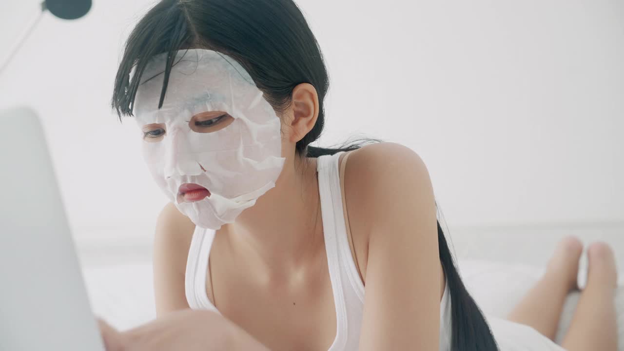 美丽的年轻亚洲女人躺在床上用单面膜在笔记本电脑上工作在卧室，美丽的女孩在家里用面膜护肤品的皱纹，皮肤护理，健康和健康。