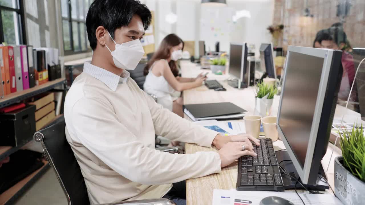 新型冠状病毒肺炎的现代办公室新常态工作环境，亚洲商人通过屏风给客户打电话，朋友做生意，戴防护口罩，保持社交距离以确保安全，中间侧视