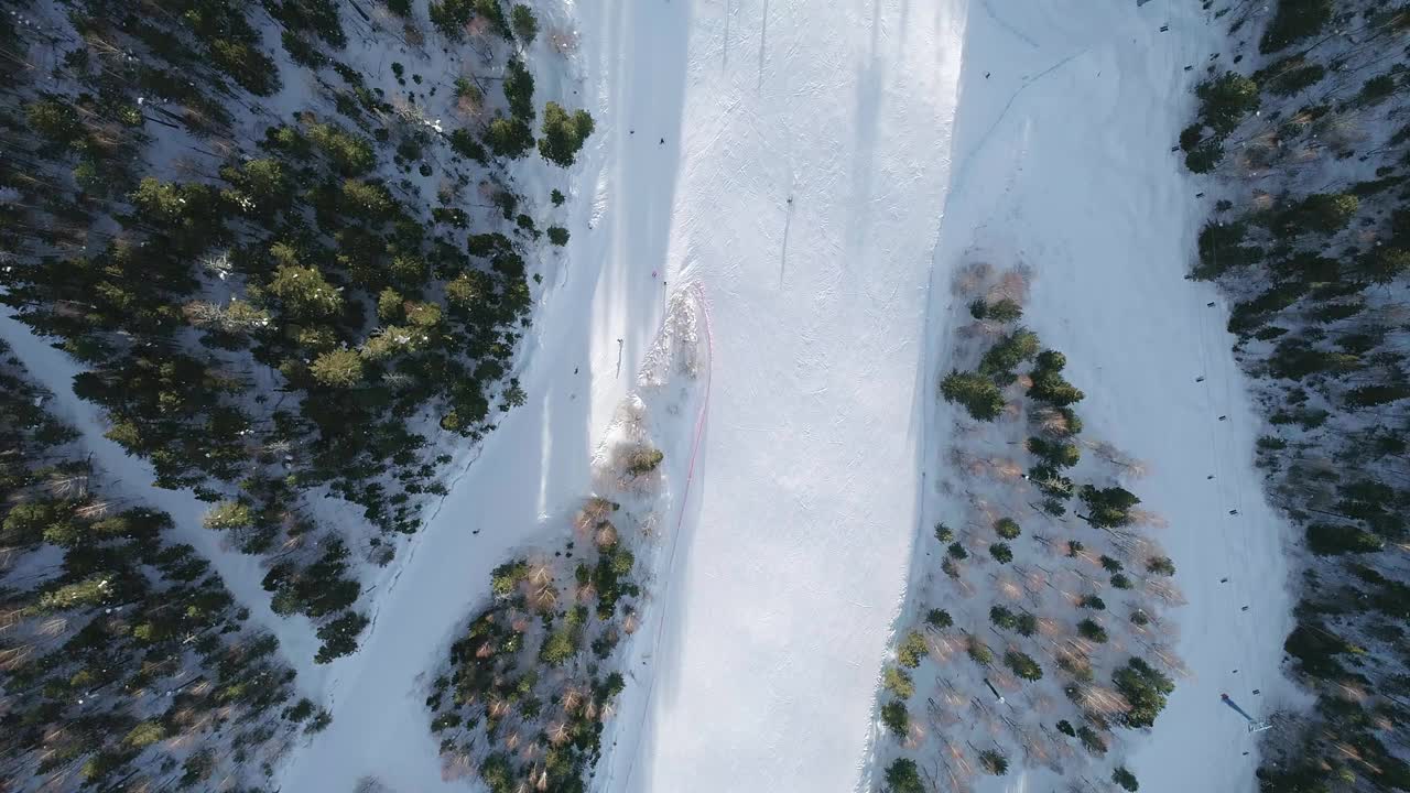 在晴朗的冬日里，滑雪者沿着树木之间的滑雪斜坡向下俯瞰，滑雪椅在一边向上移动。