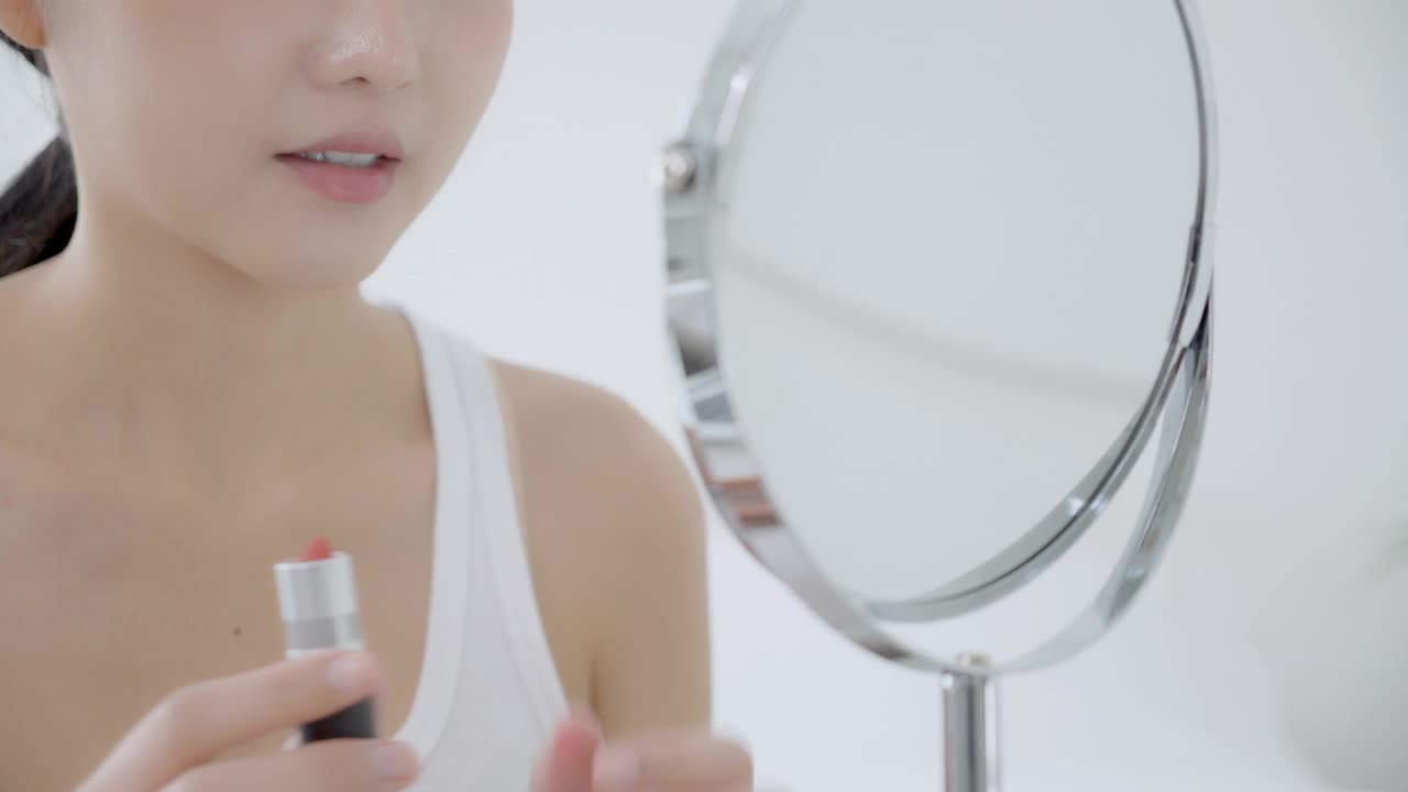 美丽的年轻亚洲女人涂口红在嘴上，美女看着镜子化妆化妆用嘴唇优雅，化妆用女性的性感，护肤和健康理念。