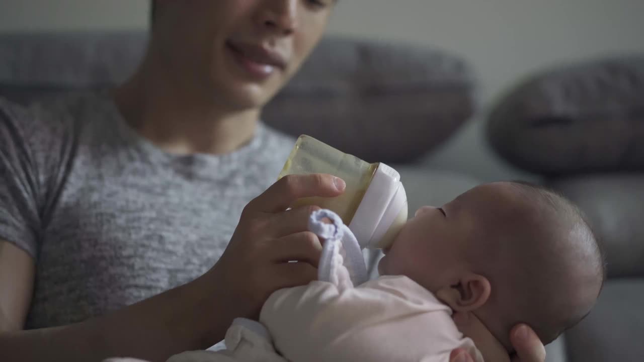 疲惫的亚洲中国年轻父亲打哈欠喂养他的儿子与奶瓶在客厅在周末