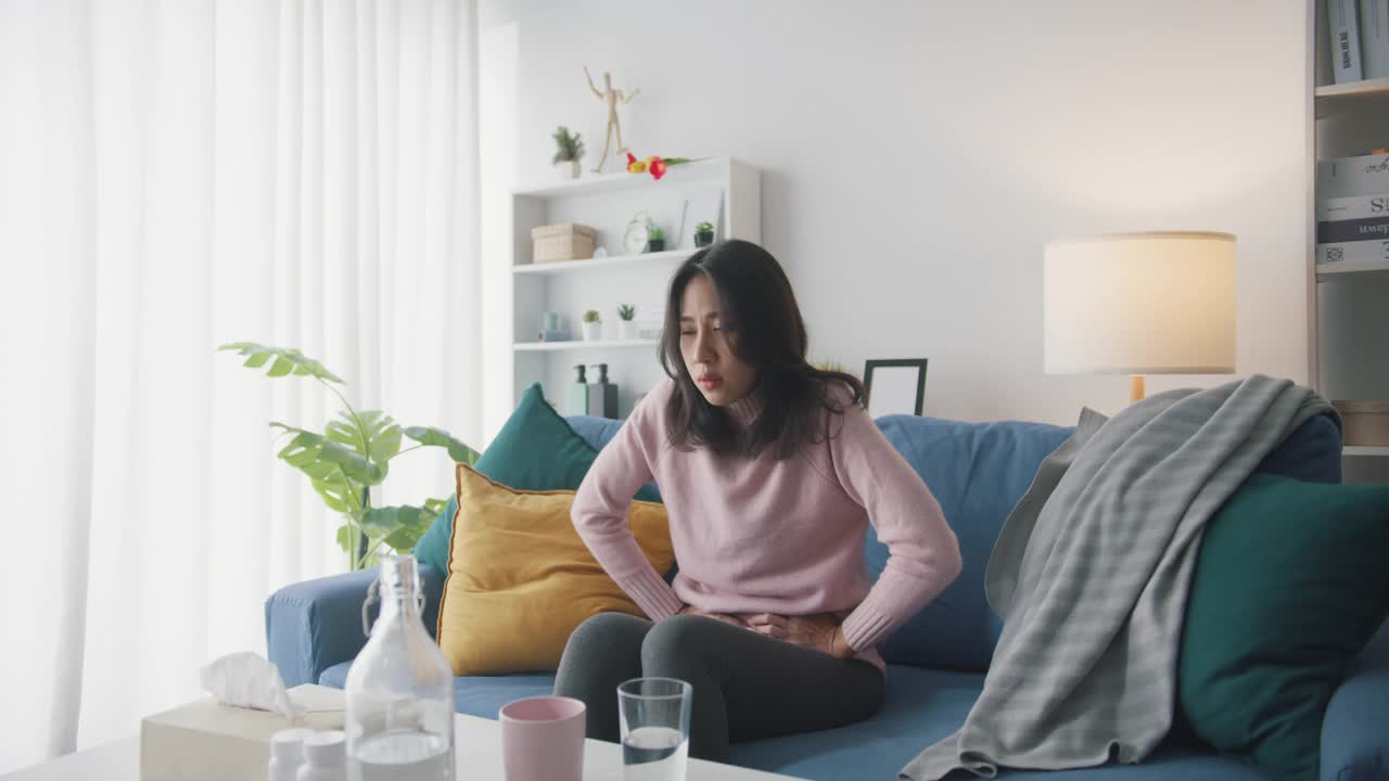 生病的亚洲妇女胃痛坐在沙发上客厅在家里。