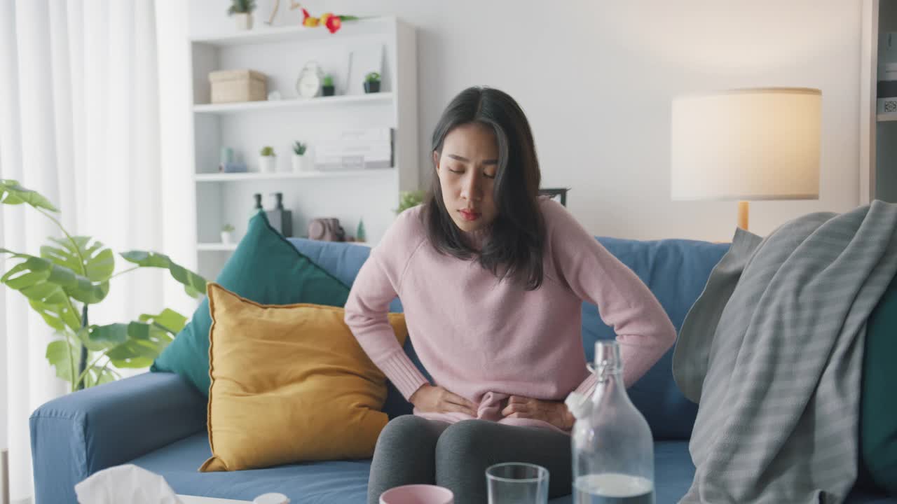 生病的亚洲妇女胃痛坐在沙发上客厅在家里。