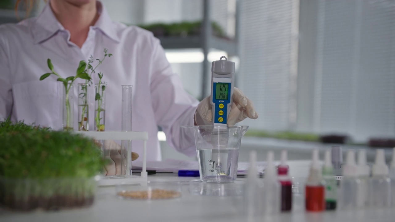 详情图-植物实验室，女实验室助理用测量设备检查水质，在现代温室的架子上灌溉年轻的微绿芽，特写镜头