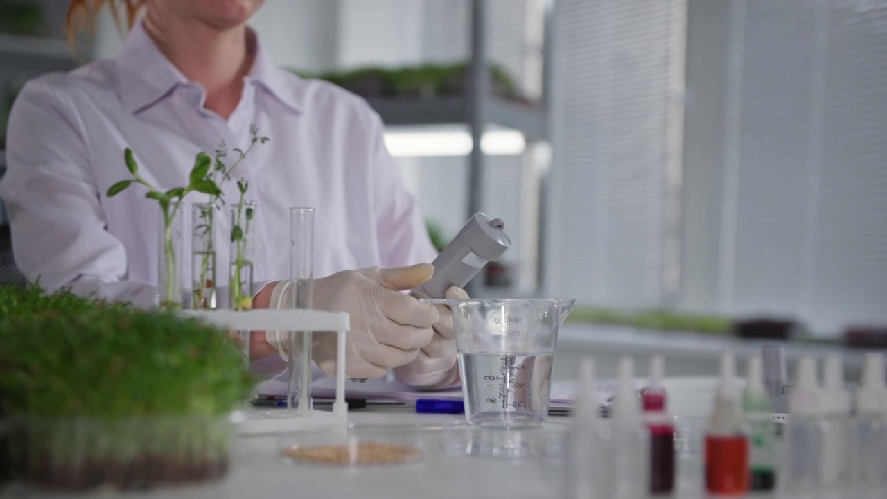 详情图-植物实验室，女实验室助理用测量设备检查水质，在现代温室的架子上灌溉年轻的微绿芽，特写镜头