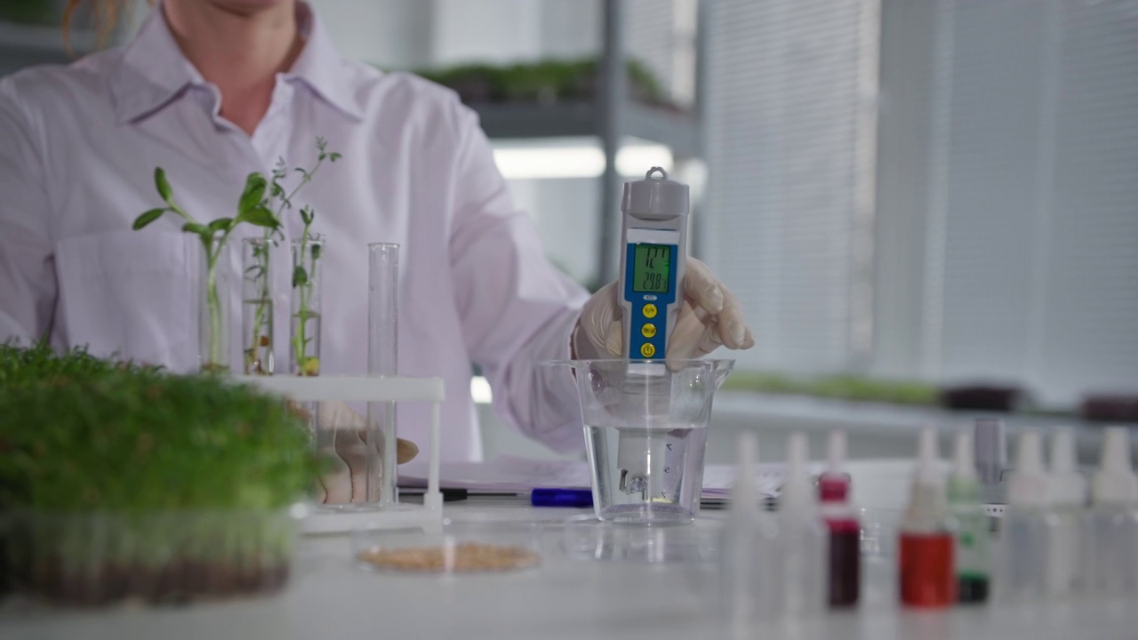 详情图-植物实验室，女实验室助理用测量设备检查水质，在现代温室的架子上灌溉年轻的微绿芽，特写镜头