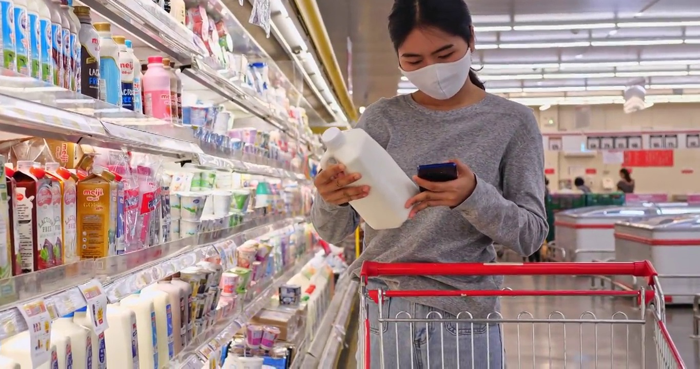 年轻的亚洲女性带着防护口罩在超市购买日用品。她正在阅读写在包装上的产品信息。