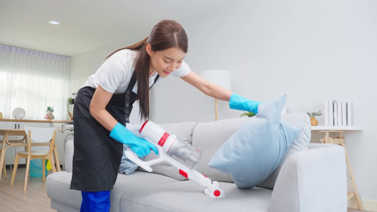 亚洲清洁服务女工清洁客厅在家里。美丽的年轻女孩管家清洁工穿围裙和吸尘肮脏的地板，家务和家务在房子。