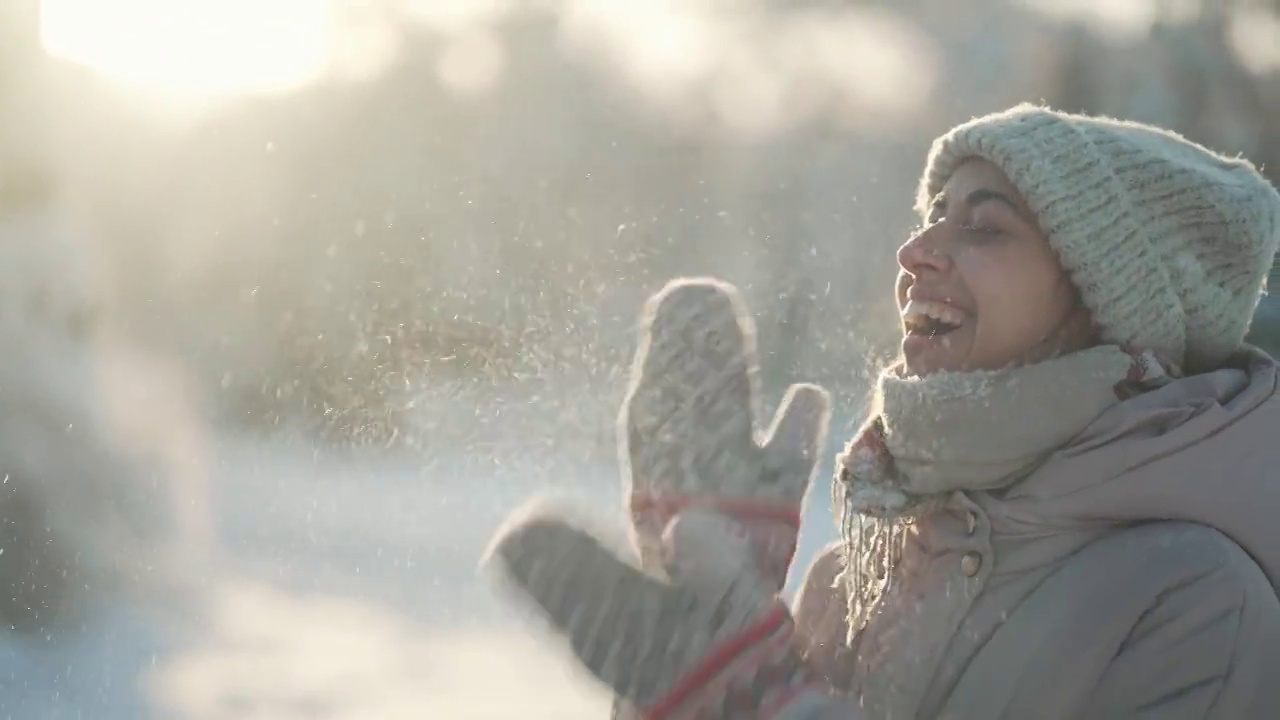 慢动作:年轻女子吹雪。在霜冻的冬日里，一个有趣的女孩在冬季公园里从手套里吹雪。快乐兴奋的女孩，享受着霜冻的新鲜，玩着新鲜的雪