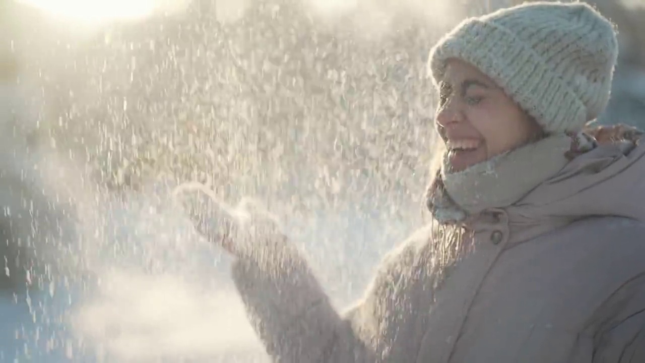 详情图-慢动作:年轻女子吹雪。在霜冻的冬日里，一个有趣的女孩在冬季公园里从手套里吹雪。快乐兴奋的女孩，享受着霜冻的新鲜，玩着新鲜的雪