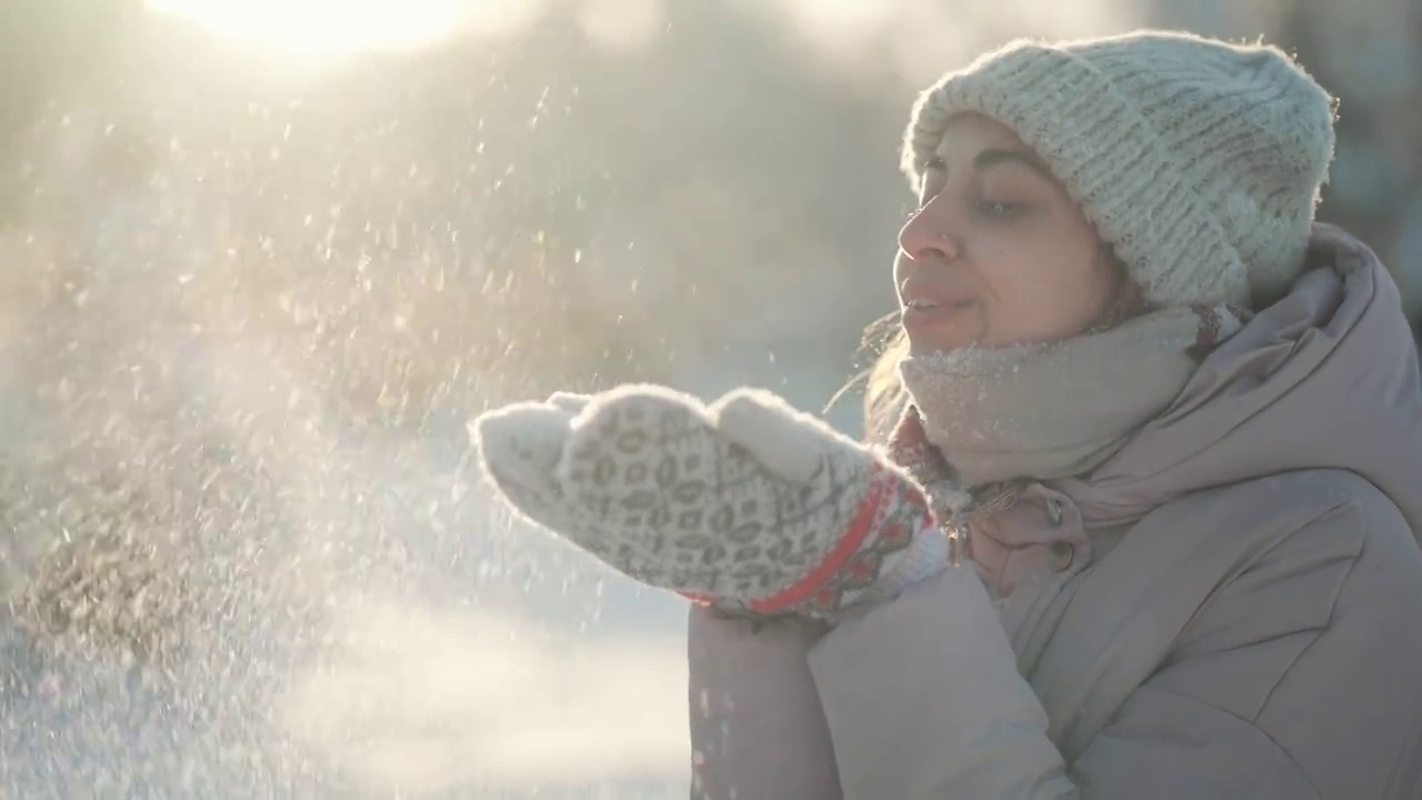 详情图-慢动作:年轻女子吹雪。在霜冻的冬日里，一个有趣的女孩在冬季公园里从手套里吹雪。快乐兴奋的女孩，享受着霜冻的新鲜，玩着新鲜的雪