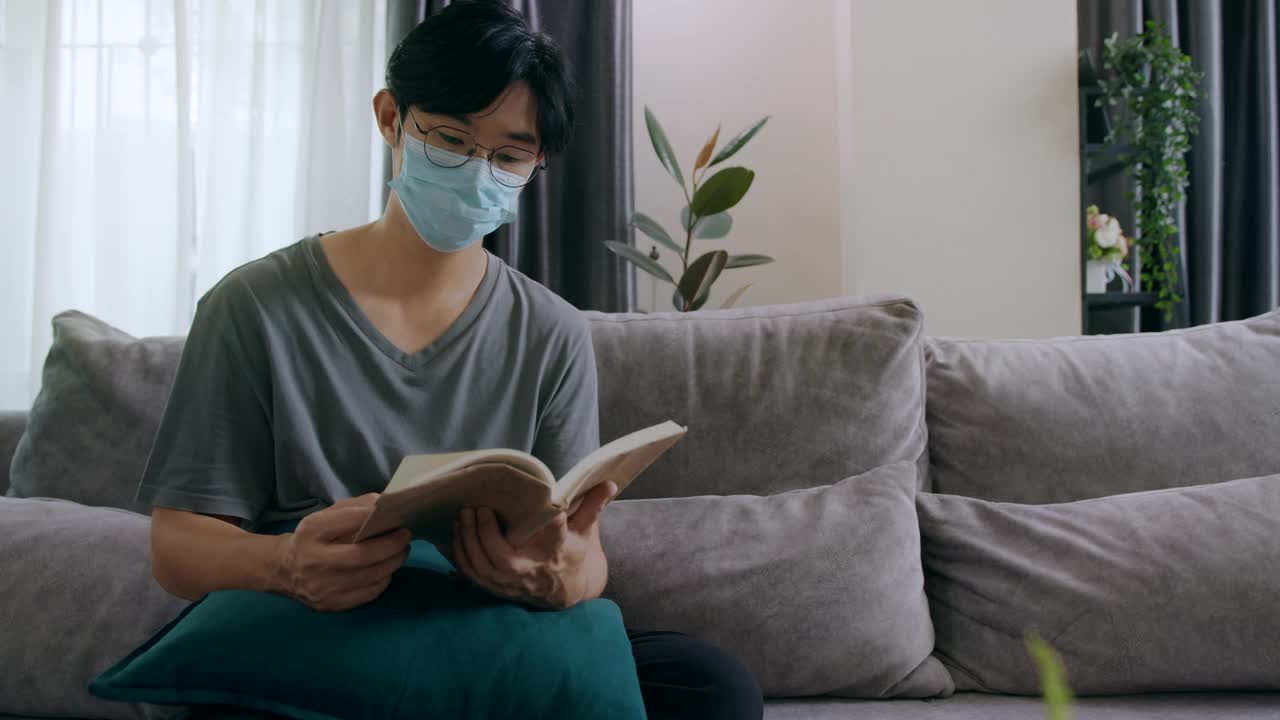 自我隔离病人隔离男子戴医用口罩在客厅看书在家隔离。保持社交距离，喉咙痛，心理健康