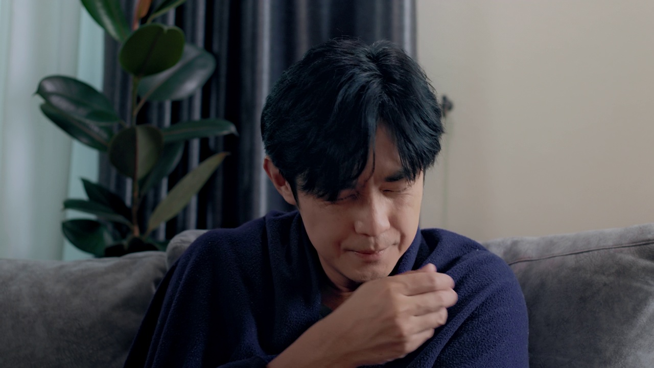 特写亚洲生病的自由职业男子与高烧坐在沙发上感觉强烈的头痛盖上毯子在家里。疾病季节性病毒问题感冒和偏头痛。