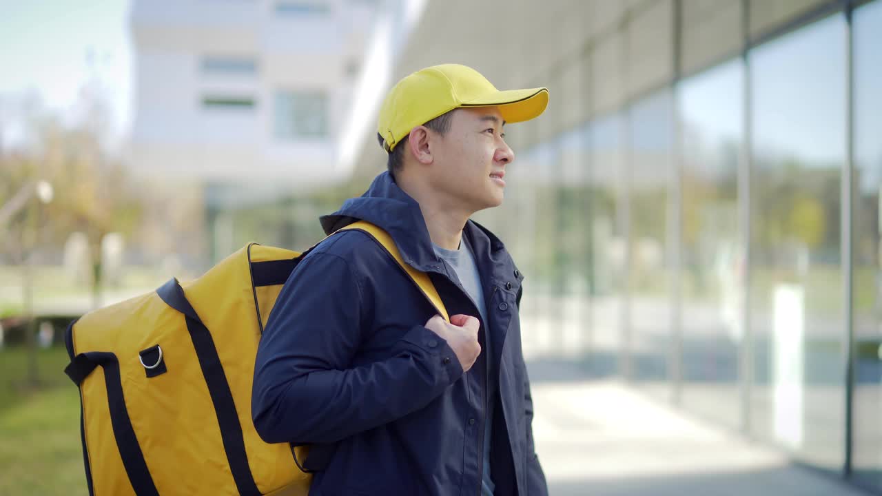 亚洲快乐男性快递服务人员的肖像，戴着帽子，背着背包。城市街道上的员工，在城市背景下看着镜头微笑