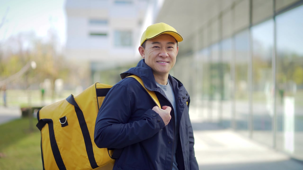 详情图-亚洲快乐男性快递服务人员的肖像,戴着帽子,背着背包。城市街道上的员工,在城市背景下看着镜头微笑