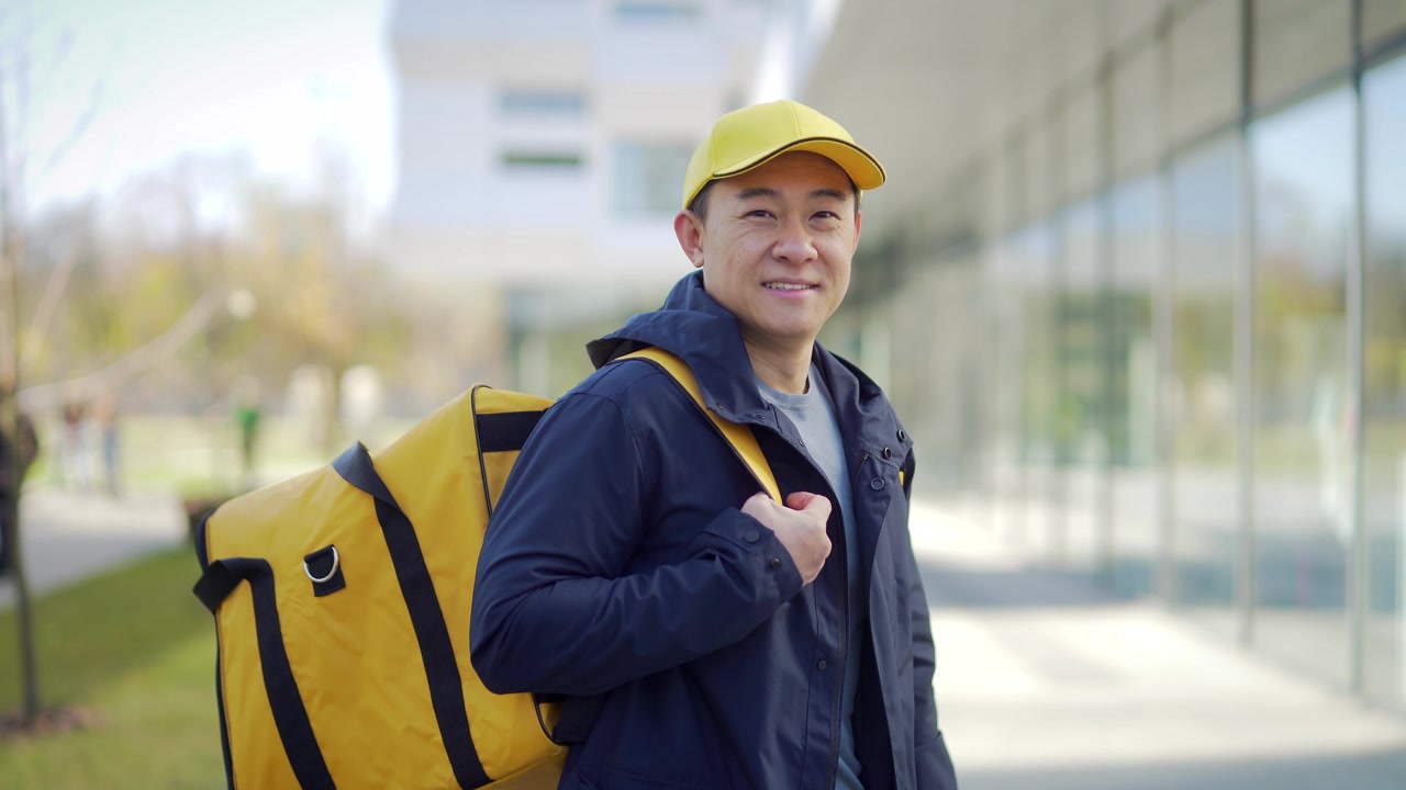 详情图-亚洲快乐男性快递服务人员的肖像,戴着帽子,背着背包。城市街道上的员工,在城市背景下看着镜头微笑