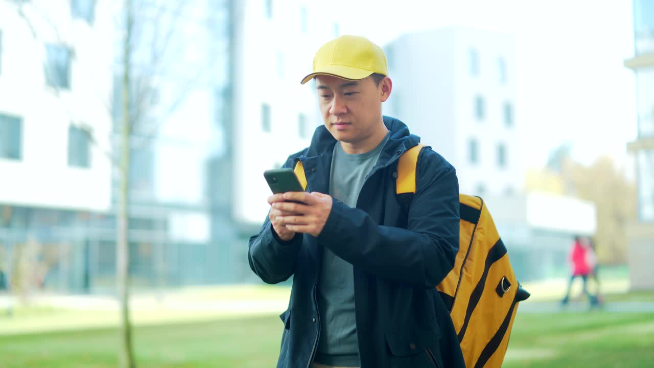 亚洲快乐男性快递服务人员的肖像，戴着帽子，背着背包。城市街道上的员工，在城市背景下看着镜头微笑。