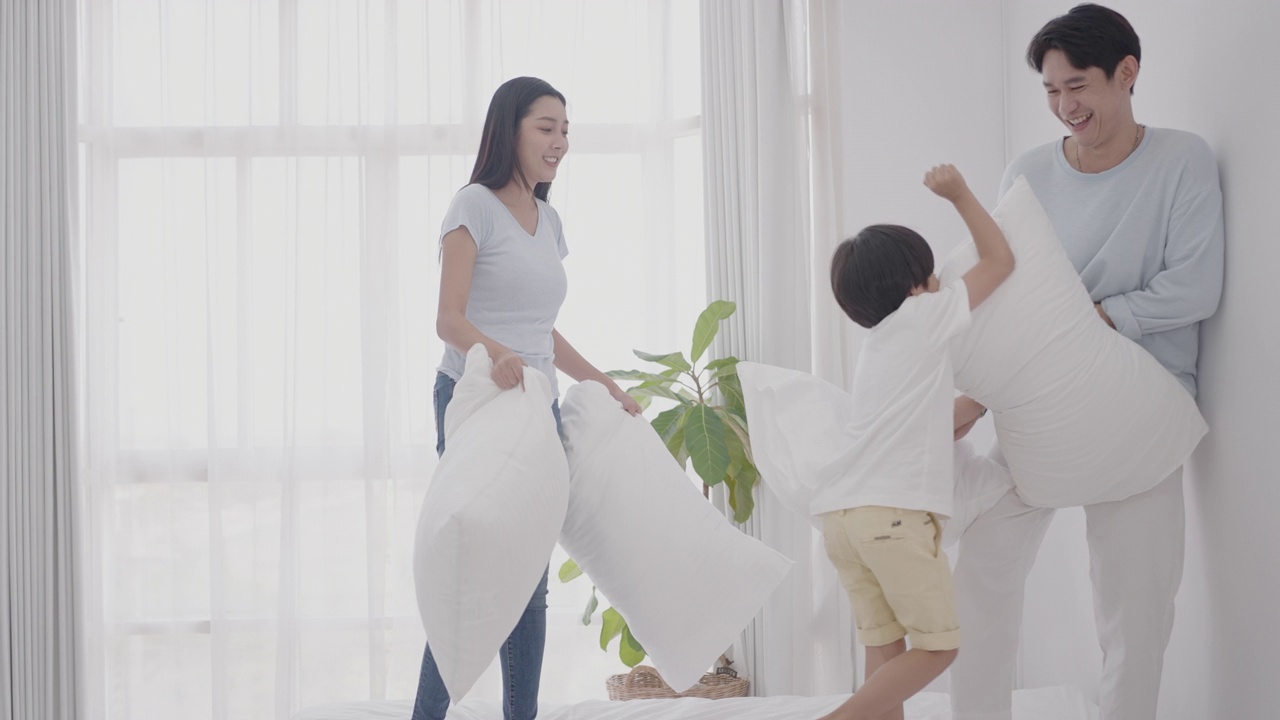 快乐的亚洲家庭在舒适的家中享受周末休闲时光。欢快的爸爸、妈妈、可爱的儿子一起在床上蹦蹦跳跳，欢天喜地，在卧室舒适的床上嬉笑打闹枕头。