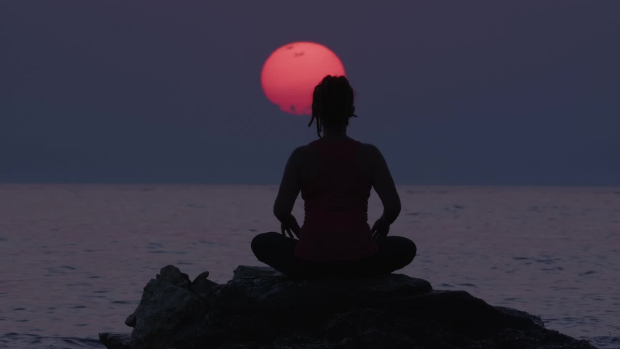 在令人惊叹的日落天空和大太阳期间，坐在海水中的石头上冥想的女人的剪影。治疗身体，心理和精神状态的人。