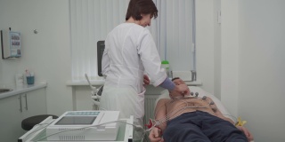 心脏病专家医生对躺在医院沙发上的一名男子进行心电图检查。现代临床男性心电图检查。诊断、保健、医疗服务。医院病人心电图检查