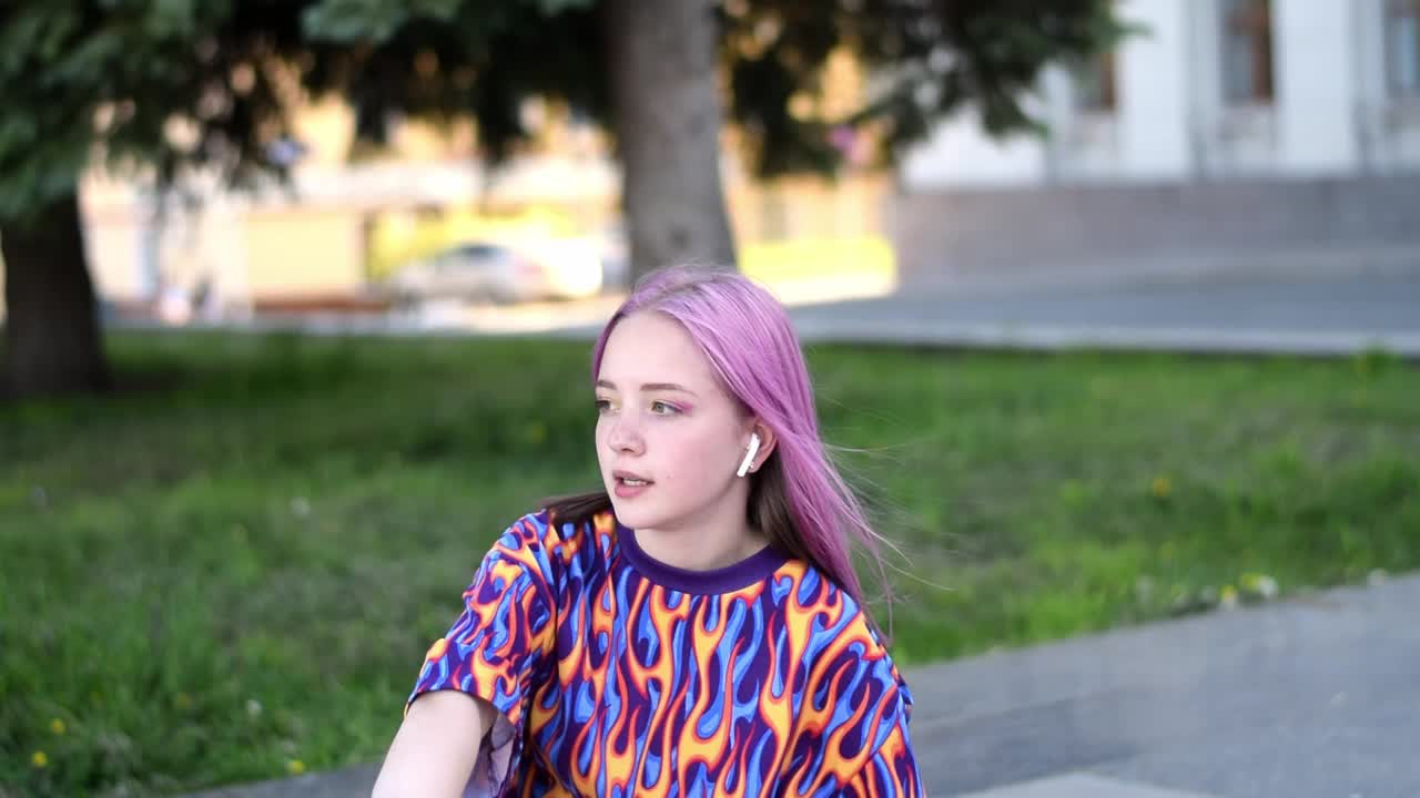 一个年轻的潮女孩，粉红色的头发，穿着彩色的t恤，戴着耳机，微笑着坐在外面的滑板上。Z一代的风格。